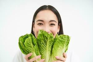 Healthy food and vegan lifestyle. Close up portrait of smiling asian woman, looks happy, shows her face with lettuce, eats cabbage, white background photo