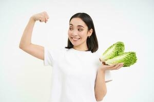 fuerte asiático mujer muestra Fresco verduras, lechuga y su músculos, flexionando bíceps con sonriente rostro, blanco antecedentes foto