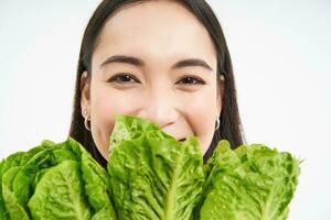 Healthy food and vegan lifestyle. Close up portrait of smiling asian woman, looks happy, shows her face with lettuce, eats cabbage, white background photo