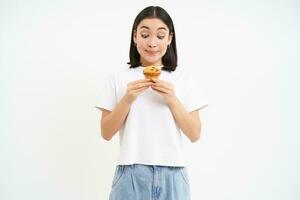 Portrait of cute girl looks at tasty cupcake, wants to bite dessert, likes pastry, white background photo