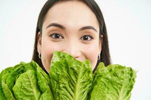 Close up portrait of happy korean woman, shows her face with lettuce, eating cabbage, likes vegetables, feels healthy and energized, white background photo