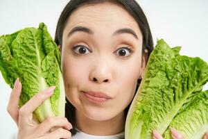 Close up portrait of young korean woman, holds lettuce next to healthy natural face without blemishes, vegetarian likes vegetables, white background photo