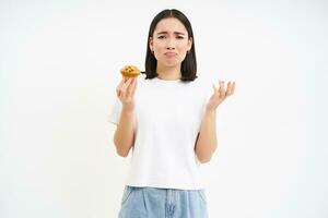 Portrait of unhappy korean woman looks at cupcake with sad face, girl on diet cant eat pastry, concept of healthy food, white background photo