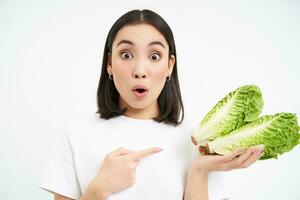 Portrait of surprised and amazed asian woman, points at cabbage, find out amazing benefit of lettuce, healthy vegetables, white background photo