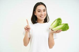 Image of smiling asian girl shows cabbage and one finger, points, teaches healthy diet, stands over white background with lettuce photo