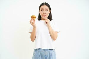 Image of young modern woman with cupcake, looks at delicious pastry from bakery with happy face, white background photo