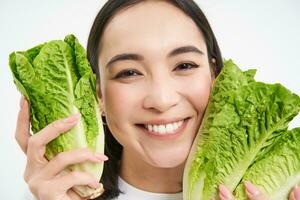 Healthy food and vegan lifestyle. Close up portrait of smiling asian woman, looks happy, shows her face with lettuce, eats cabbage, white background photo