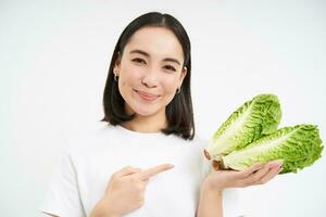 Smiling asian woman, pointing finger at cabbage, grew fresh vegetable, looks happy, isolated on white background photo
