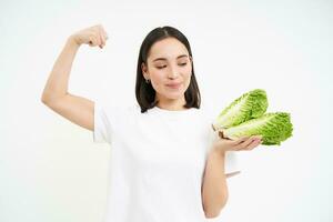 Strong asian woman shows fresh vegetables, lettuce and her muscles, flexing biceps with smiling face, white background photo