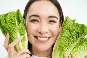 Healthy food and vegan lifestyle. Close up portrait of smiling asian woman, looks happy, shows her face with lettuce, eats cabbage, white background photo