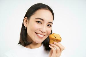 Close up portrait of asian woman likes pastry, bites tasty cupcake with happy unbothered smile, white background photo