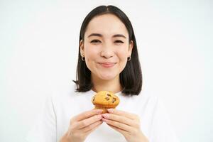 Close up portrait of asian woman likes pastry, bites tasty cupcake with happy unbothered smile, white background photo