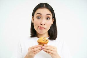 Close up portrait of asian woman with thoughtful face, looks at cupcake, wants to bite dessert, thinks of calories, white background photo