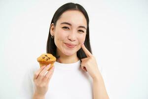 Close up portrait of smiling korean woman, eating cupcake with pleased face, white background photo