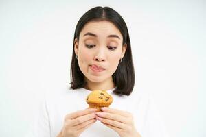 Close up portrait of asian woman with thoughtful face, looks at cupcake, wants to bite dessert, thinks of calories, white background photo