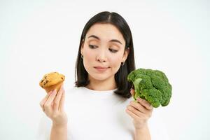 Nutrition and healthy diet. Smiling korean woman, holding broccoli and cupcake, choosing between vegetables and pastry, white background photo