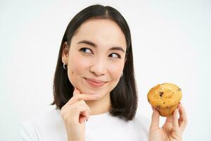 Close up portrait of korean woman with one cupcake, thinking and smiling, white studio background photo