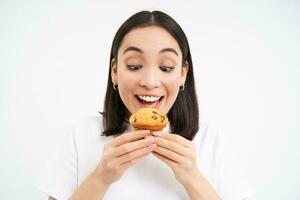 Close up portrait of asian woman likes pastry, bites tasty cupcake with happy unbothered smile, white background photo