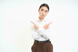 Puzzled businesswoman points sideways, shows two choices, makes decision, stands over white background photo