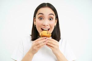 Close up portrait of asian woman likes pastry, bites tasty cupcake with happy unbothered smile, white background photo