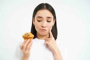 Image of pensive korean woman, looks at cupcake with thoughtful face, thinking about dessert, white background photo