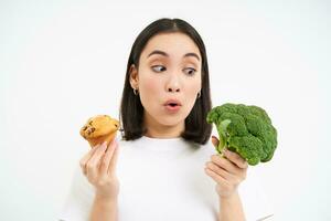 Portrait of asian woman looks excited at broccoli, holding cupcake, white background photo