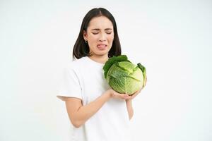 asiático mujer mira con asco y disgusto a repollo, odia verduras, aislado en blanco antecedentes foto