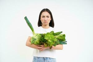 Unhappy korean woman with vegetables, staying on diet with raw food, frowning upset, white background photo