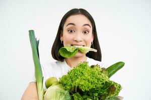 cerca arriba retrato de asiático niña masticando lechuga hoja, participación verde oránico verduras, Guías sano estilo de vida con nutritivo alimento, blanco antecedentes foto