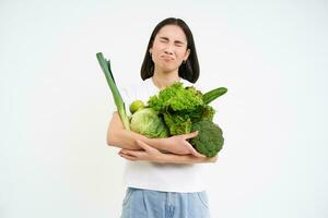Miserable asian woman, holds green vegetables and complains at het diet, looks at calories, white background photo
