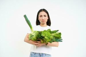 Image of sad, unhappy girl on diet, holding vegetables, green raw food, complaining, standing over white background photo