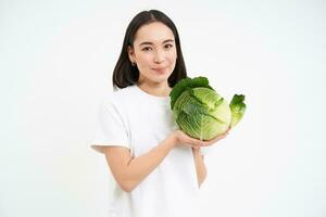 retrato de encantador coreano mujer, demostración repollo, participación lechuga verde vegetal y sonriente, aislado en blanco antecedentes foto