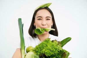 retrato de sonriente coreano mujer come lechuga hoja, participación verde verduras, comiendo nutritivo alimento, blanco antecedentes foto