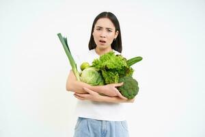 Sad and gloomy asian woman, holding vegetables, organic food for detor and nutrition, white studio background photo