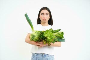 Image of sad, unhappy girl on diet, holding vegetables, green raw food, complaining, standing over white background photo