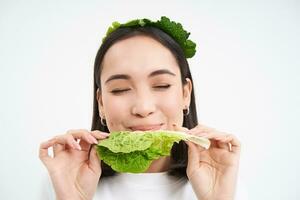 feliz, linda asiático mujer comiendo verde lechuga, vegetariano gustos oránico verduras, blanco estudio antecedentes foto