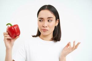 Portrait of asian woman looking at red sweet pepper with reluctant, dislike face, white background photo