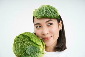 Close up portrait of beautiful and healthy vegan woman, holding lettuce, has cabbage lead on head, white background photo