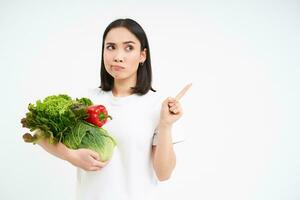 Portrait of pensive asian woman, holding vegetables, pointing right with thinking face, white background photo