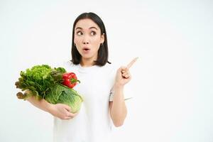 Portrait of girl with surprised face, pointing at advertisement, holding green vegetables, concept of dietry, white background photo