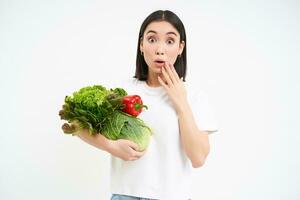 Image of girl with vegetables, looks surprised at camera, isolated on white background photo