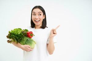 Enthusiastic korean woman, holding bunch of vegetables, pointing finger right at banner, showing promo advertisement, white background photo