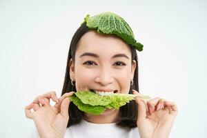 retrato de linda sonriente coreano mujer con hoja en cabeza, come repollo y mira feliz, blanco antecedentes foto