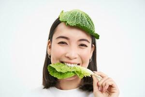 retrato de asiático niña con hoja en cabeza, come repollo y sonrisas, aislado en blanco antecedentes foto