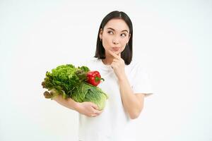 imagen de pensativo asiático mujer con verduras, pensando y mirando aparte, Cocinando verde orgánico alimento, blanco antecedentes foto