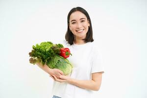 retrato de sonriente sano mujer, vegetariano muestra verde orgánico vegetales y mirando feliz, aislado en blanco antecedentes foto