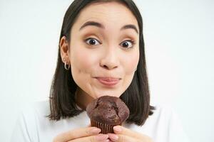 cerca arriba de linda sonriente asiático mujer, participación chocolate magdalena cerca boca, teniendo morder, disfruta comiendo Pastelería, blanco antecedentes foto