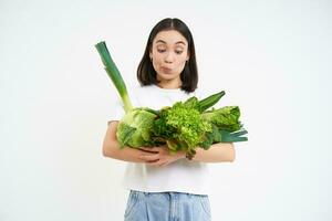 Healthy vegan lifestyle. Happy asian woman looks at her favourite vegetables, holding cabbages, lettuce and green organic food, white background photo