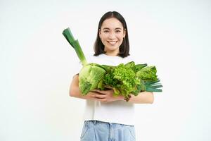 imagen de sonriente asiático mujer, vegetariano con un montón de verde verduras, comiendo oránico alimento, aislado en blanco antecedentes foto
