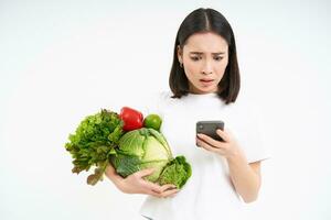 Portrait of asian woman looks worries at smartphone screen, holds vegetables, orders food online, white background photo